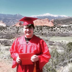 A young man with disabilities in a red graduation gown standing in front of a mountain, symbolizing his independence as a young adult.