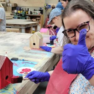 A woman is learning and developing real-world skills by making a birdhouse in a workshop, while potentially developing friendships with fellow participants.