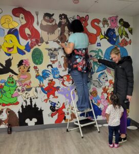 At Zach's Place, an employee assists a woman and a child in painting a mural filled with Disney characters on the wall.