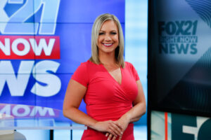 A smiling female news anchor in a red dress stands in a TV studio with "Fox21 Night of Comedy" visible in the background.