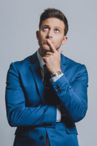 Man in blue suit looking thoughtful with a finger on his chin, gazing upward against a gray background, perhaps contemplating his next comedy night routine.