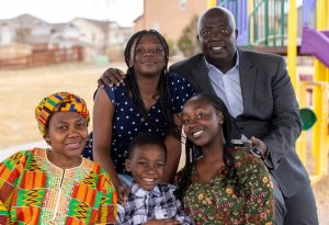 A family of five poses for a photo at a playground, where smiles abound among adults and children alike. The woman seated on the left wears colorful traditional attire, reflecting their rich heritage. Behind them, houses stand as a testament to their close-knit community fostering strong family bonds.