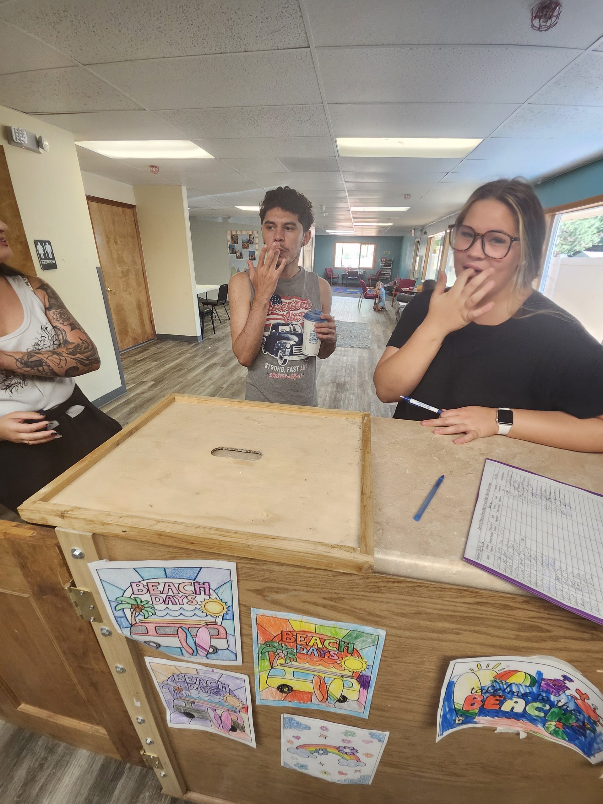 BEES_June2025(4) Three people stand at a counter in a classroom; two are making a gesture with their hands near their mouths. Coloring pages are displayed on the wooden counter.