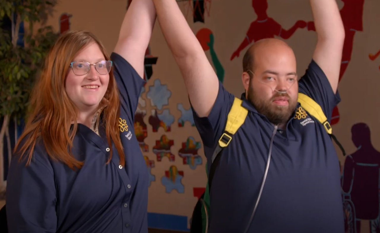CommunityImmunity(1) Two adults wearing navy shirts with yellow logos sit side by side, smiling and raising one arm each in celebration. A colorful wall mural is visible in the background.