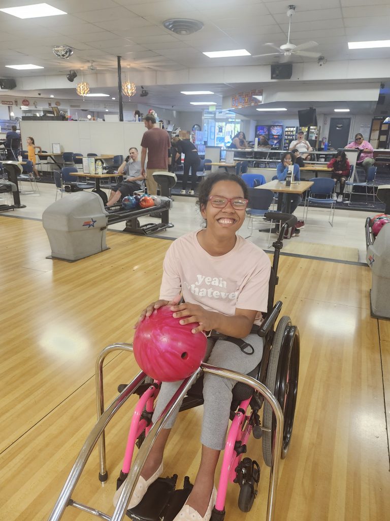 20240918_180936 A smiling person in a wheelchair holds a pink bowling ball on a bowling lane, with people and bowling equipment visible in the background.