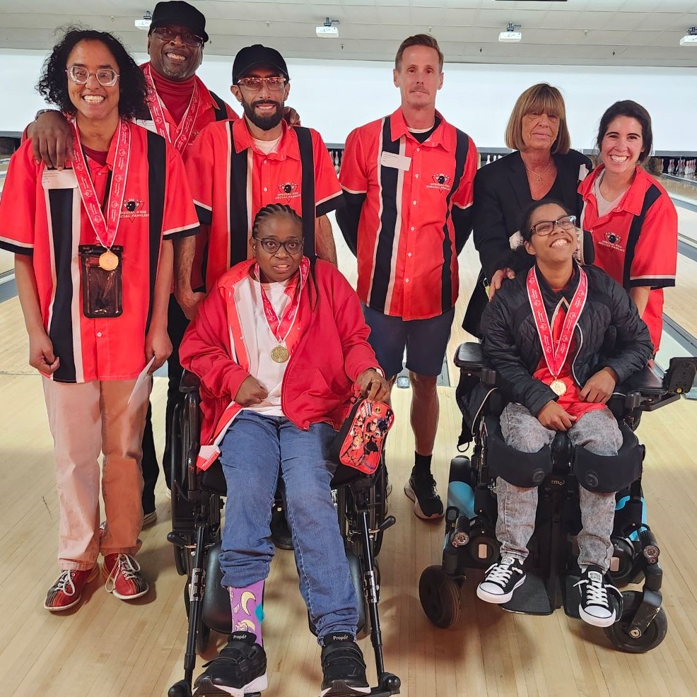 image000001 A group of eight people, some in wheelchairs, pose together on a bowling lane wearing matching red-and-black shirts and medals around their necks.