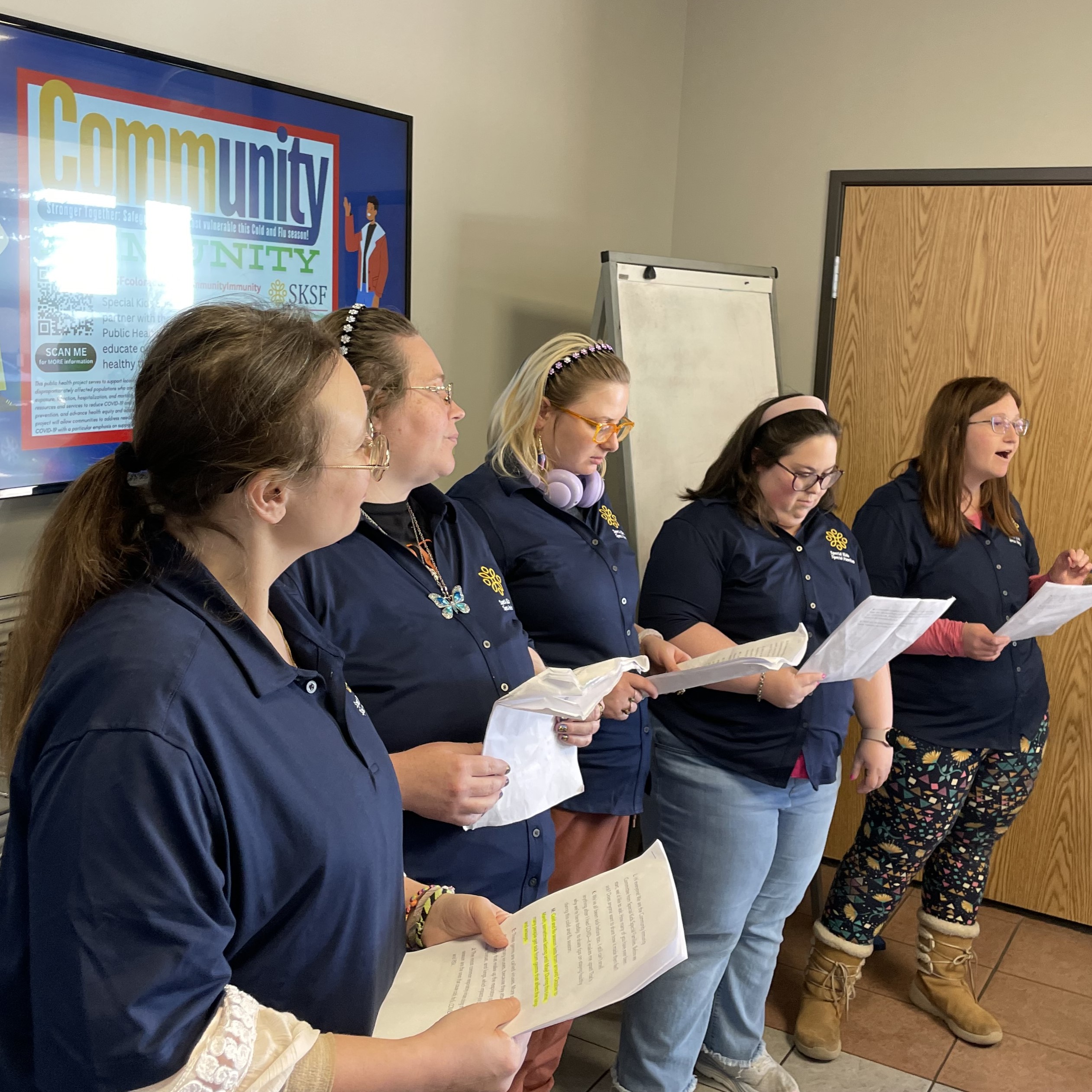 CIC_Feb.Mar2025-6 Five women wearing matching navy blue shirts stand in a row indoors, holding papers and singing or speaking, with a "Community" poster visible in the background.