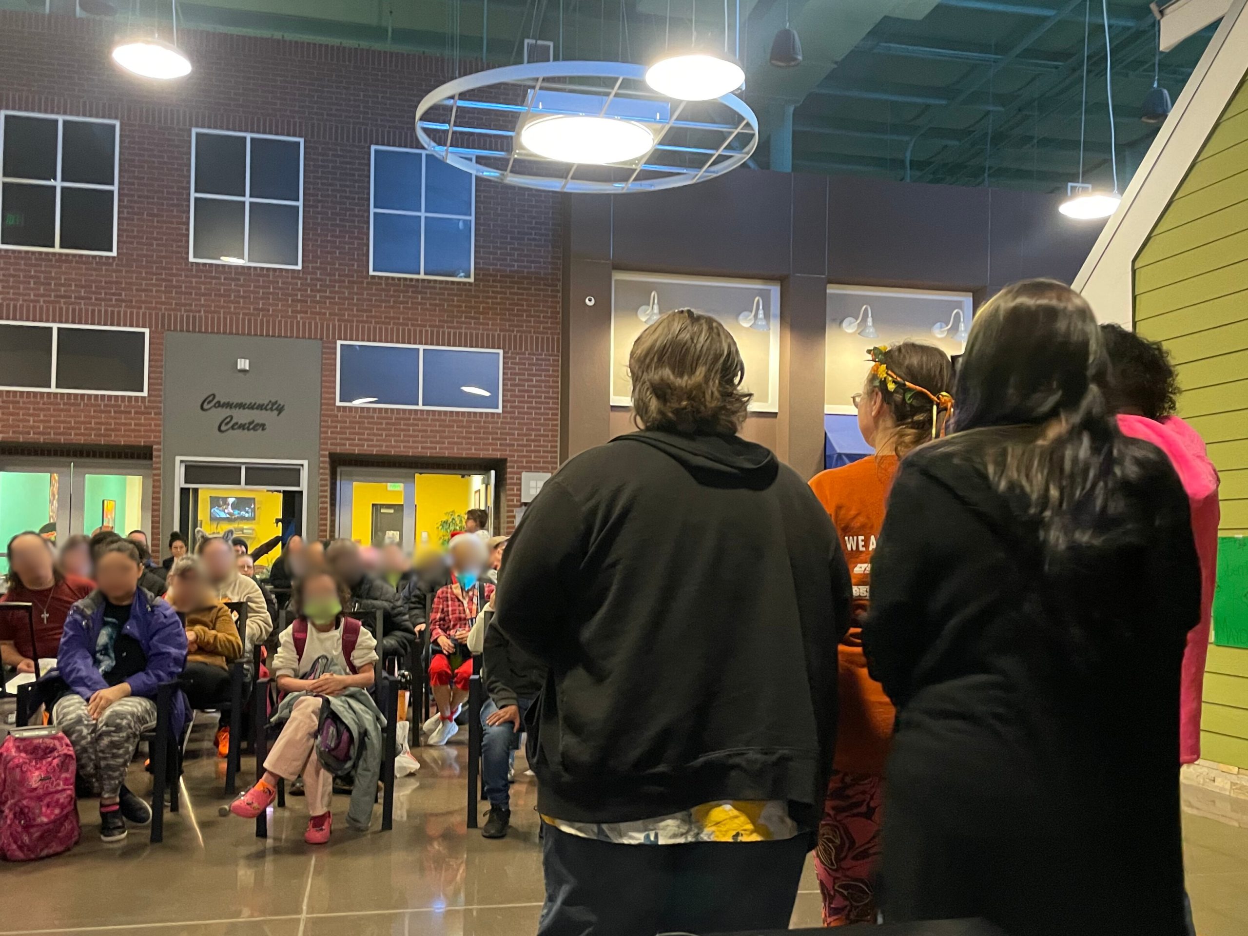 COMMUNITY.IMMUNITY_atGoodwill_Nov2024(2) A group of people stands facing an audience seated in a community center with brick walls and overhead lighting.