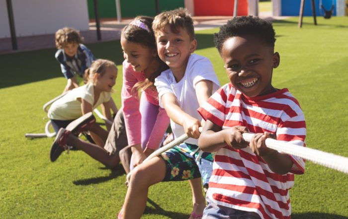 Five children play tug-of-war on a grassy playground, smiling and pulling a rope, with colorful walls and playground equipment in the background.