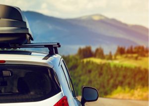 A white car with a roof box is parked on a road, overlooking a scenic landscape near Colorado Springs, with green hills and distant mountains under a partly cloudy sky.