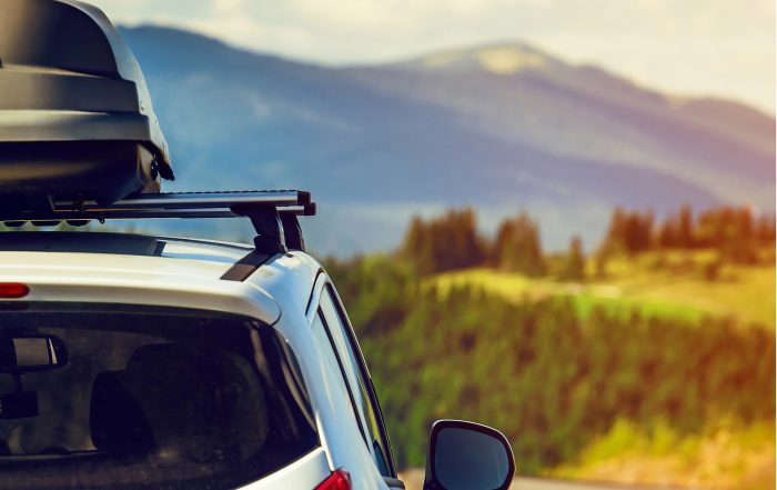 A white car with a roof box is parked on a road, overlooking a scenic landscape near Colorado Springs, with green hills and distant mountains under a partly cloudy sky.