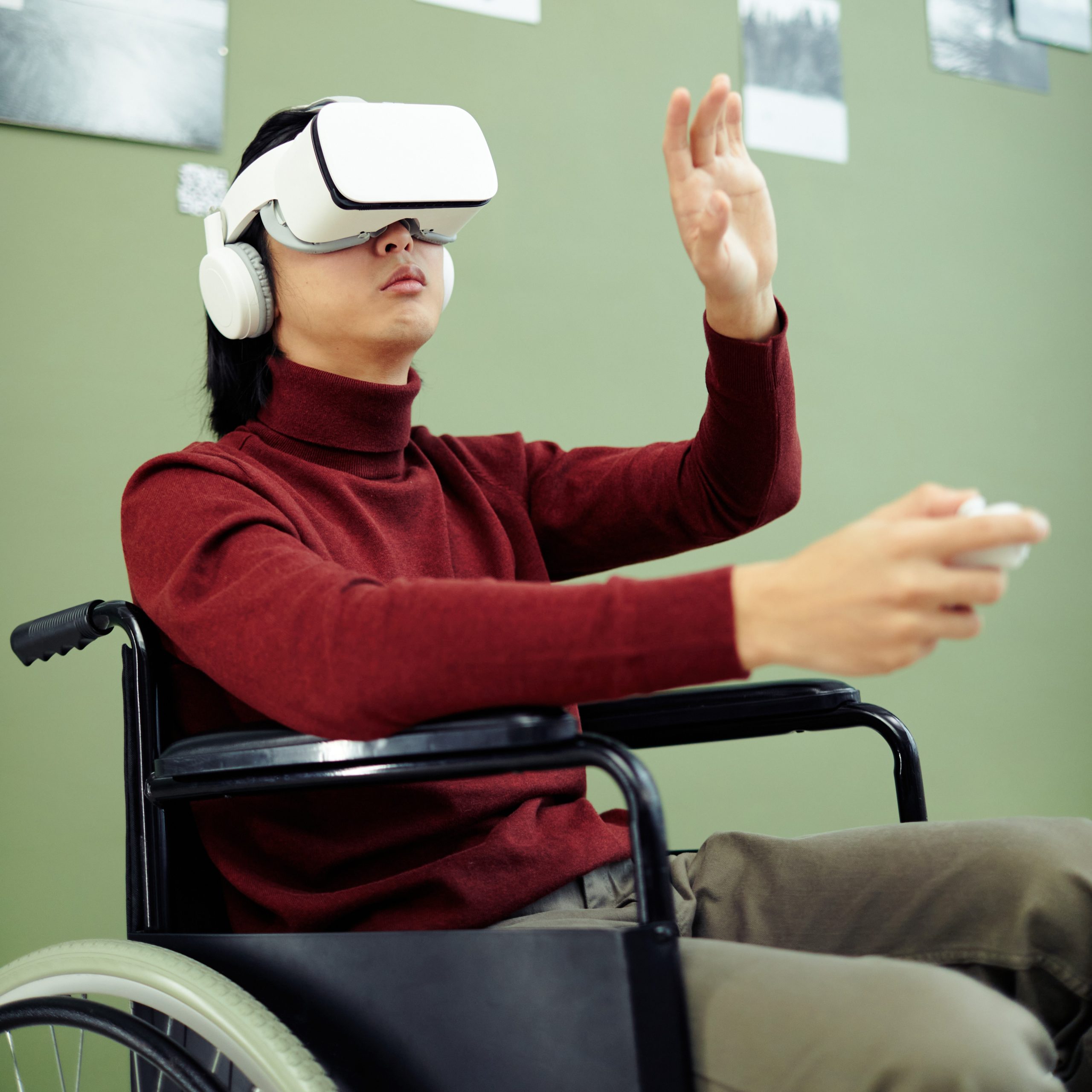 A person in a wheelchair uses virtual reality goggles and headphones, holding a controller and raising one hand, against a green background with photos on the wall.
