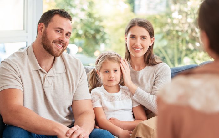A man, woman, and young girl sit together on a couch, smiling and listening to another person whose back is to the camera.