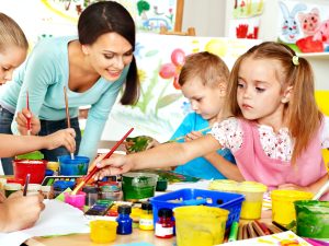 A teacher supervises young children painting and drawing at a table filled with art supplies, fostering child development and kindergarten readiness in a vibrant classroom setting.