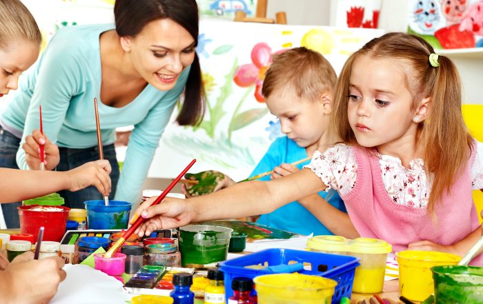 A teacher supervises young children painting and drawing at a table filled with art supplies, fostering child development and kindergarten readiness in a vibrant classroom setting.