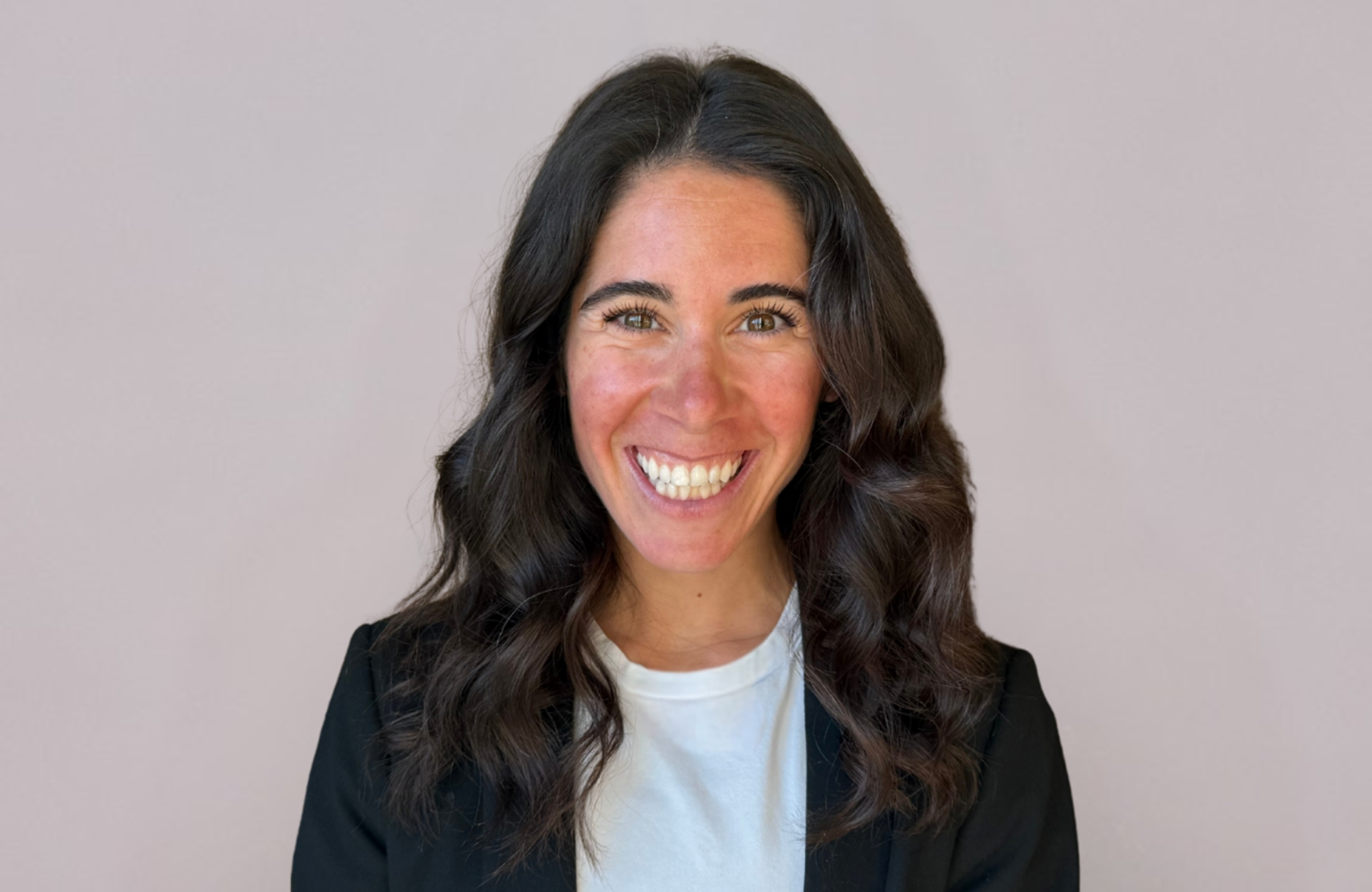 Woman with long, wavy dark hair smiling at the camera, wearing a white shirt and black blazer against a plain light background.