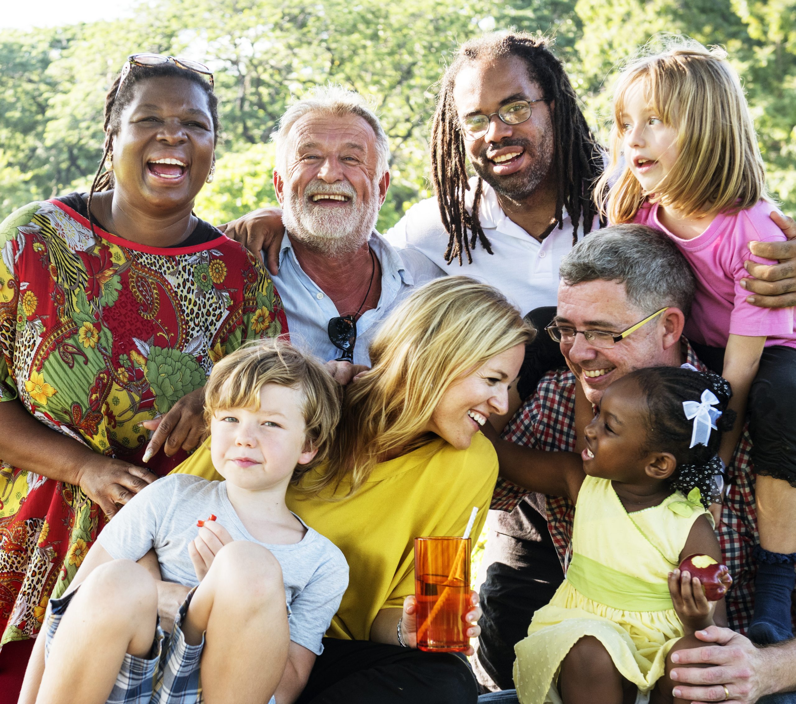 A group of adults and children sit closely together outdoors, smiling and laughing, with trees in the background.