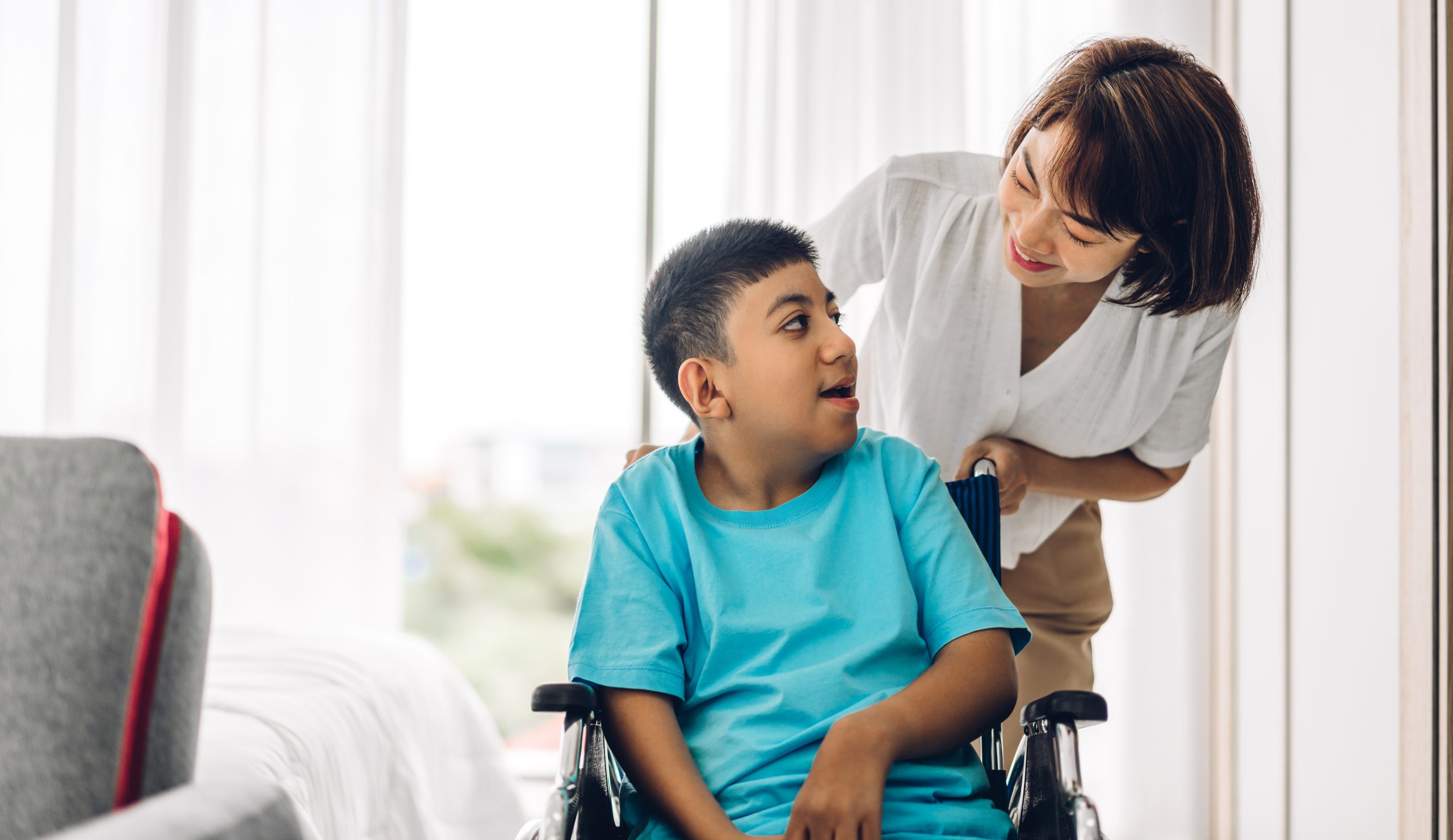 A woman stands behind a young boy in a wheelchair. They are indoors, near a bed, and looking at each other, both appearing calm and engaged.